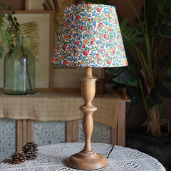 Decorative lamp with floral shade on a wooden base, placed on a table with plants in the background.