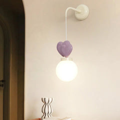 A wall sconce with a white mounting bracket, a white cord, three purple heart-shaped accents, and a white globe light emitting warm light.