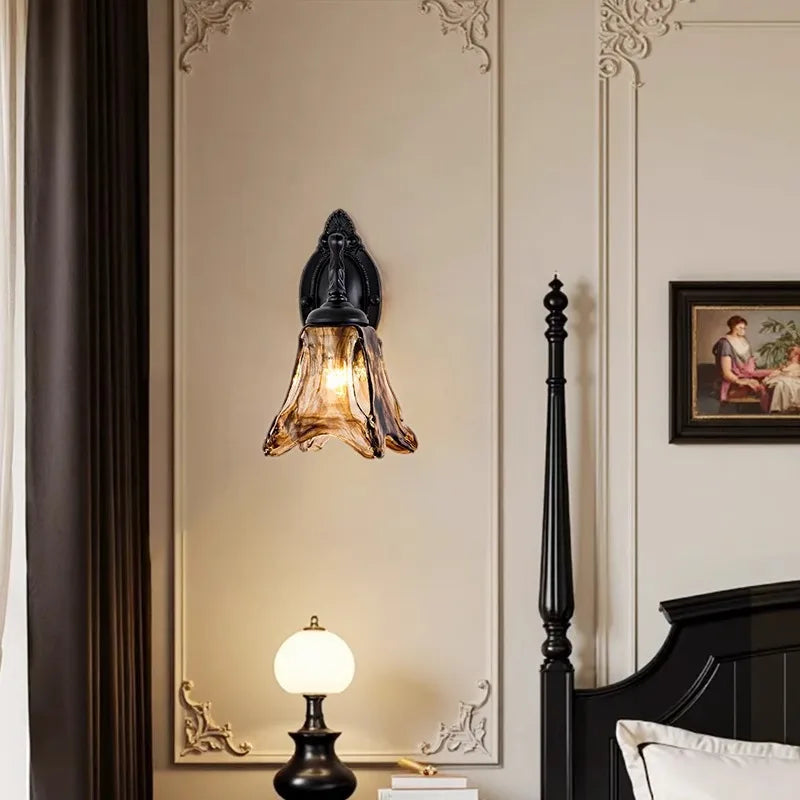 A bronze glass bell wall sconce mounted on a wall next to a dark wood bed frame, with a bedside lamp below.