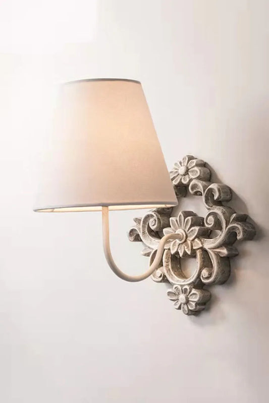 Gray white wooden wall light with a conical fabric shade and ornate floral base mounted on a white wall.