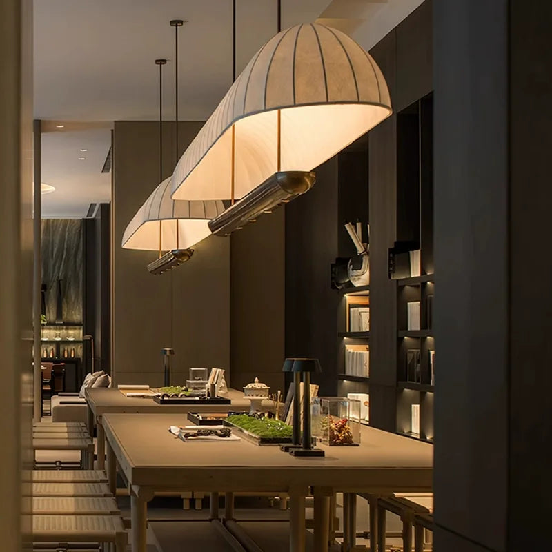 Three fabric tube pendant lights, each with a warm glow, suspended above long dining tables in a dimly lit restaurant.