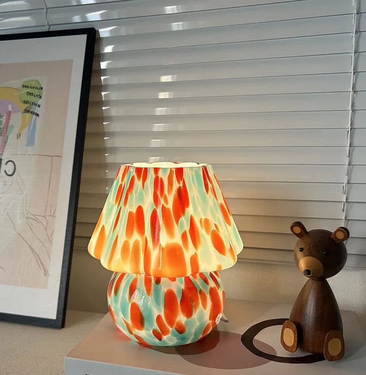 A single mushroom-shaped table lamp with a stained glass shade featuring orange and teal splatters, placed on a book next to a wooden bear figurine.