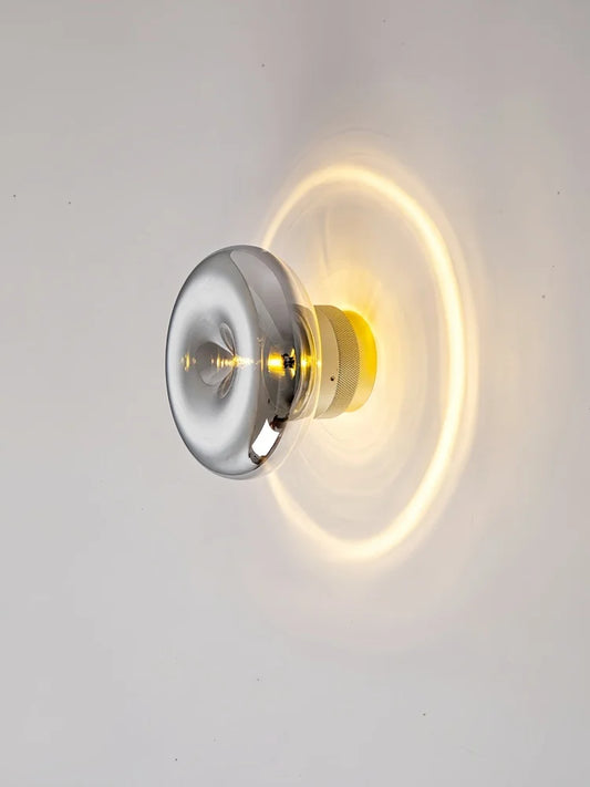 Close-up of a modern wall sconce with a smoky gray glass diffuser emitting a warm, circular halo of light on a white wall.