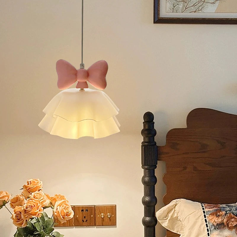 Pink bow tiered chandelier hanging above a bed, with bedside flowers and a lamp.