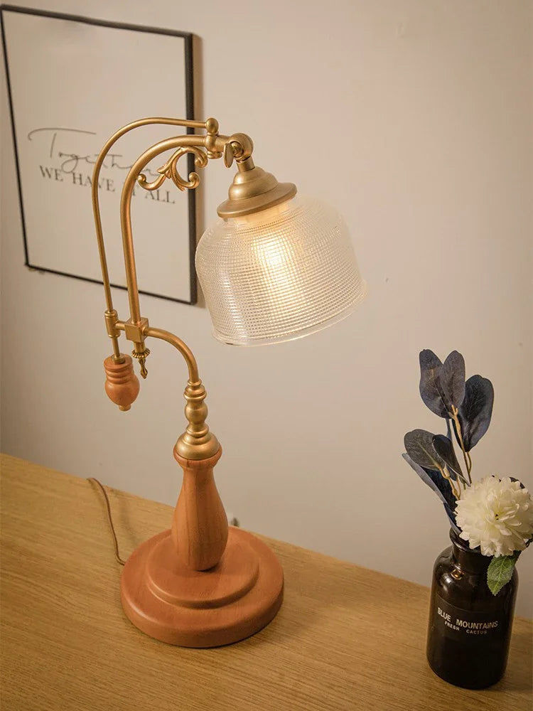 A wooden base table lamp with a frosted glass shade and brass arm, positioned on a table near a framed picture and a vase of flowers.