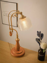 A wooden base table lamp with a frosted glass shade and brass arm, positioned on a table near a framed picture and a vase of flowers.
