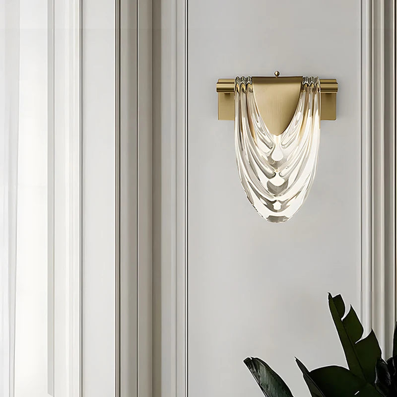 A gold wall sconce with a draped clear glass shade, mounted on a vertically striped wall.