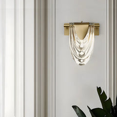A gold wall sconce with a draped clear glass shade, mounted on a vertically striped wall.
