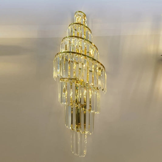 A tall, spiraling gold wall sconce with multiple tiers of clear crystals hanging from gold bands, illuminated against a light-colored wall.