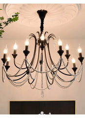 A black, multi-arm chandelier with pearl bead draping and crystal details, installed on a white ceiling with decorative molding, illuminated and casting a warm glow.