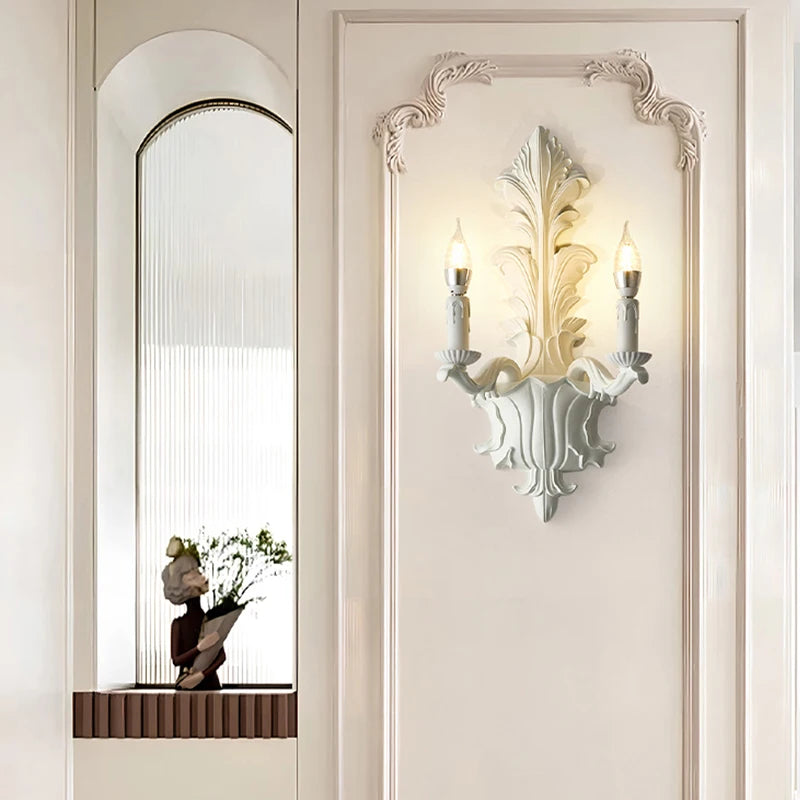 White leaf-shaped wall sconce with two candle-style lights mounted on a paneled wall, near an arched niche with a statue.