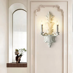 White leaf-shaped wall sconce with two candle-style lights mounted on a paneled wall, near an arched niche with a statue.