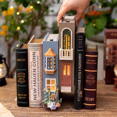 A hand placing a brown bookend lamp with an illuminated window and balcony into a row of decorative bookends on a wooden shelf.