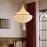 A white fabric pleated pendant light with a black trim, hanging from a white ceiling in a room with a painting on the wall and a red curtain.