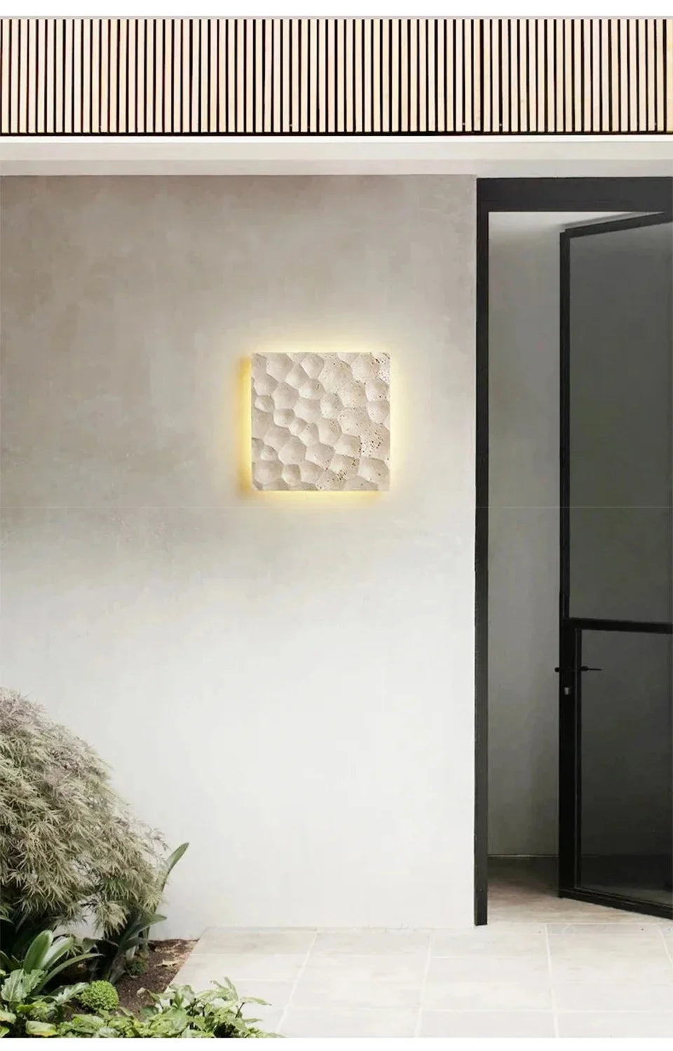A light stone textured wall sconce mounted on an exterior wall near a modern doorway and lush greenery.