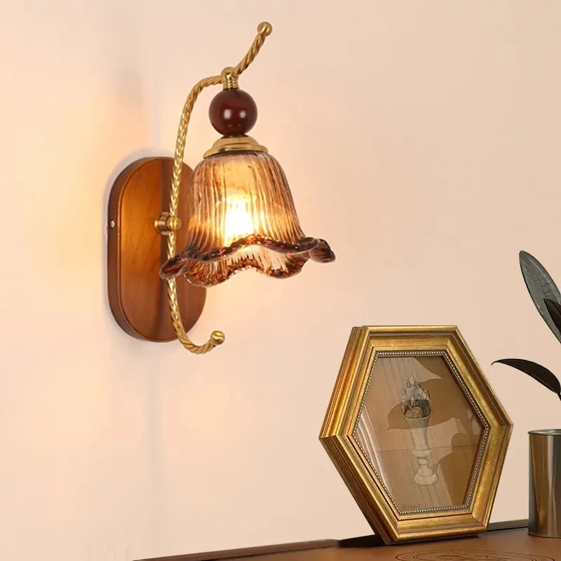 A vintage-style wall sconce with a floral-shaped amber glass shade and a gold arm mounted on a wooden oval backplate.