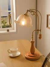 A vintage-style table lamp with a wooden base and an ornate brass arm supporting a textured glass shade, placed on a wooden table next to a teacup.