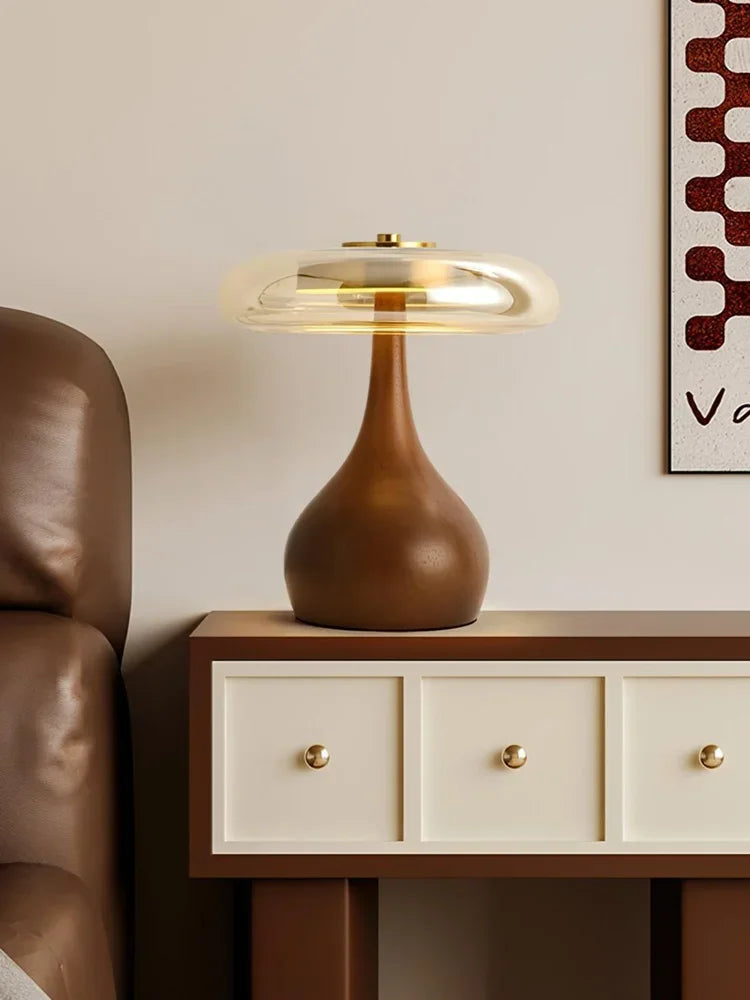 Brown table lamp on a wooden side table with a beige wall and brown leather chair in the background.