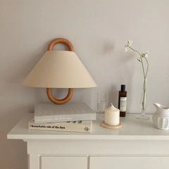 A beige fabric cone shade wall sconce with a natural wood hoop frame mounted on a wall above a shelf with books and decor.