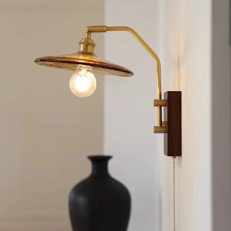 Wall-mounted lamp with a gold arm and wooden base, illuminated, next to a black vase on a neutral background.
