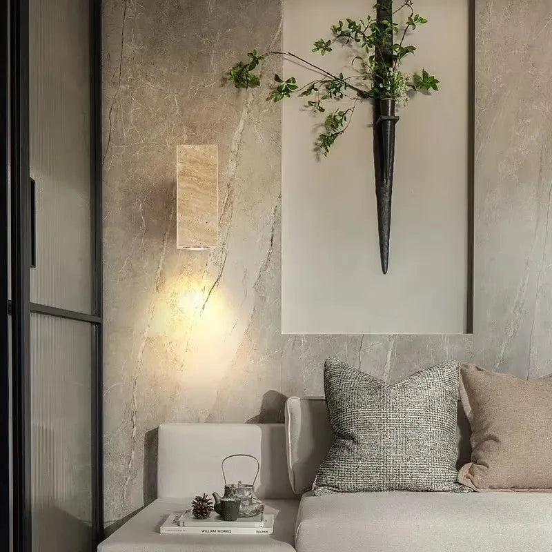 A close-up of a stone wall sconce mounted on a marble-patterned wall, with a sprig of greenery in a dark vase mounted above it.