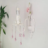 A white wall sconce with pink crystal droplets and a single LED bulb against a white wall, next to a green potted plant.