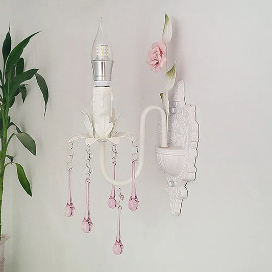 A white wall sconce with pink crystal droplets and a single LED bulb against a white wall, next to a green potted plant.