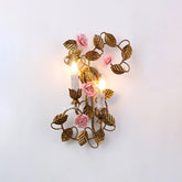 Gold leaf wall sconce with pink ceramic roses and two candle-style bulbs illuminated against a white background.