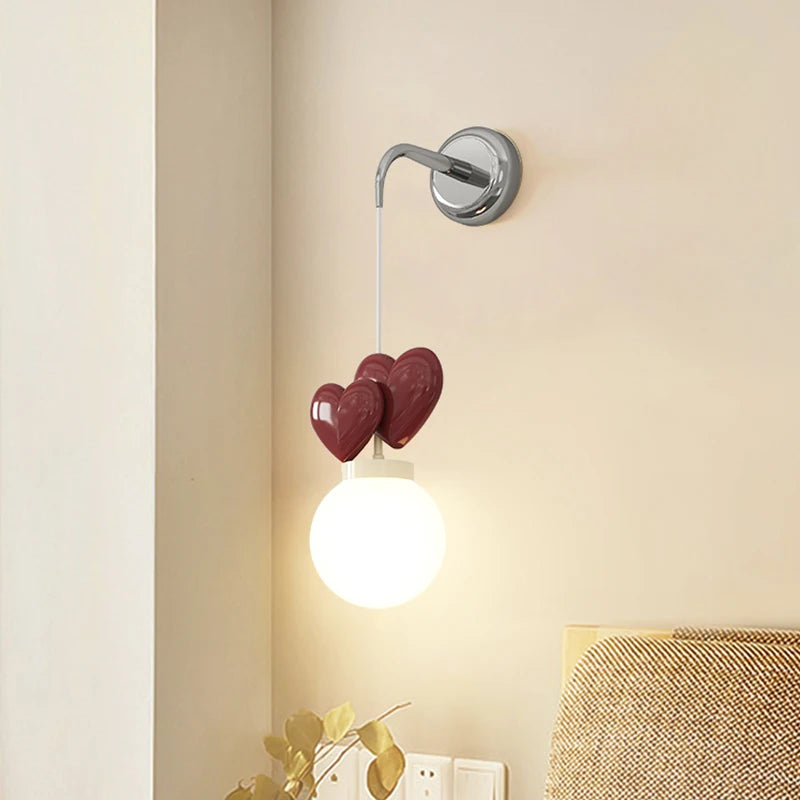 A wall sconce with a chrome mounting bracket, a white cord, three dark red heart-shaped accents, and a white globe light emitting warm light.