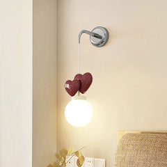 A wall sconce with a chrome mounting bracket, a white cord, three dark red heart-shaped accents, and a white globe light emitting warm light.