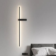 A black minimalist LED strip wall sconce with warm lighting above a grey upholstered headboard.