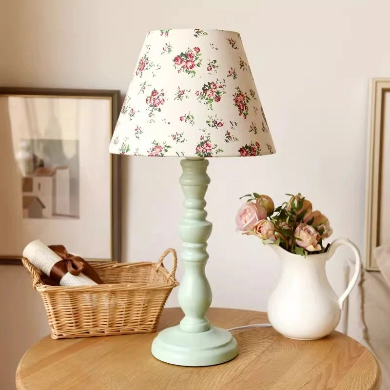 Light green table lamp with floral-patterned shade on a wooden table, next to a basket and vase with flowers.