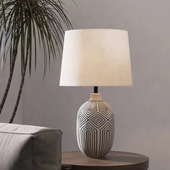 A ceramic table lamp with a patterned base and a cream fabric shade, placed on a wooden side table next to a sofa and a palm plant.