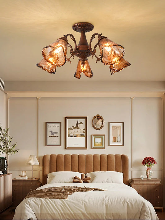 A semi-flush mount ceiling light with a brushed bronze finish and five amber stained glass shades arranged in a floral pattern, illuminating a bedroom.