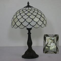 A Tiffany-style table lamp with a white and black stained glass dome shade and a dark iron base, shown on a side table next to a framed photo.