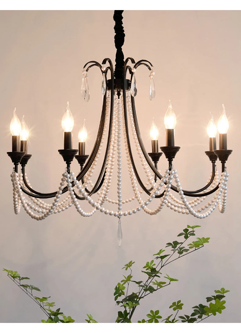 View of a black, multi-arm chandelier with pearl beads and crystal accents, partially framed by green foliage against a light-colored wall.