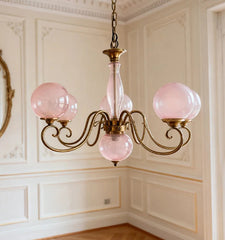Close-up of a gold pendant light with pink glass globes and ornate arms, mounted in a room with ornate wall paneling.