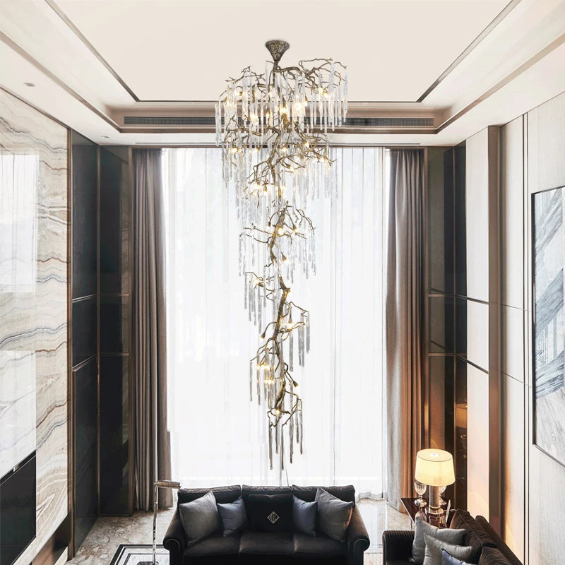 A tall, cascading chandelier with a gold branch-like frame adorned with numerous clear crystal icicle-like droplets, illuminated and hanging in a grand, high-ceiling living room.