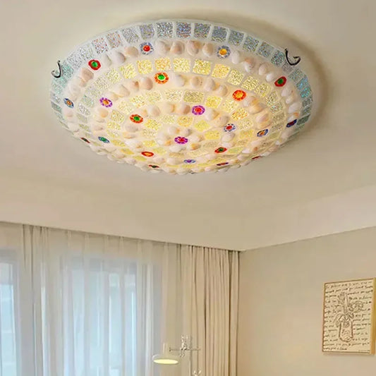 A close-up view of a round, mosaic-style flush mount ceiling light with colorful glass and shell pieces, illuminating a room.