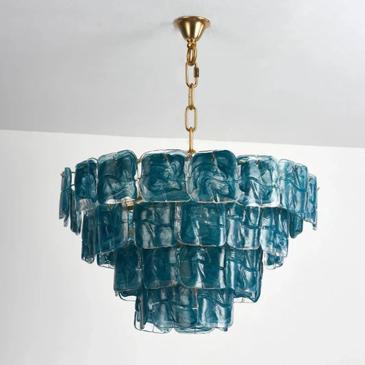 A multi-tiered chandelier with square, textured blue glass panels suspended from a gold chain and frame, against a white ceiling and wall.
