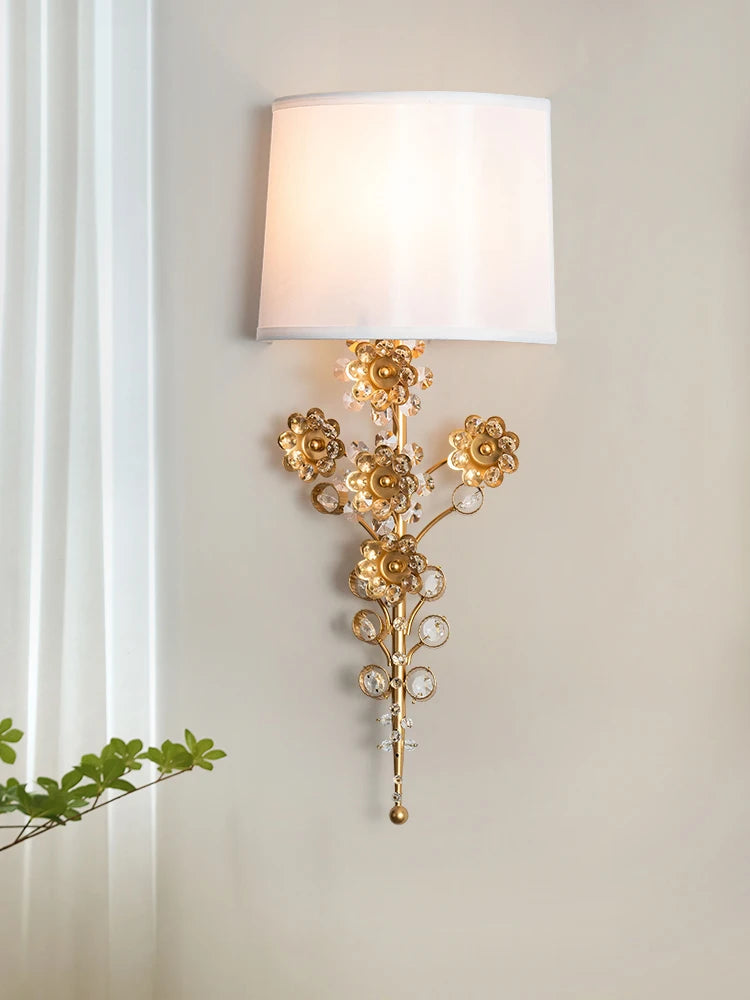 A gold floral crystal wall sconce with a white fabric shade, positioned next to a window with sheer curtains.