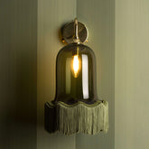 A wall-mounted brass sconce with a dark green bell-shaped glass shade and a fringe of green tassels hanging from the rim, illuminated by an incandescent bulb, against a textured green wall.