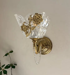 A gold wall sconce with a faceted clear glass shade and a decorative floral metal piece, mounted on a light-colored wall.