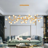 A long, horizontal gold chandelier with a branch-like design and numerous clear, fruit-shaped glass globes hangs above a light green sofa in a modern living room.