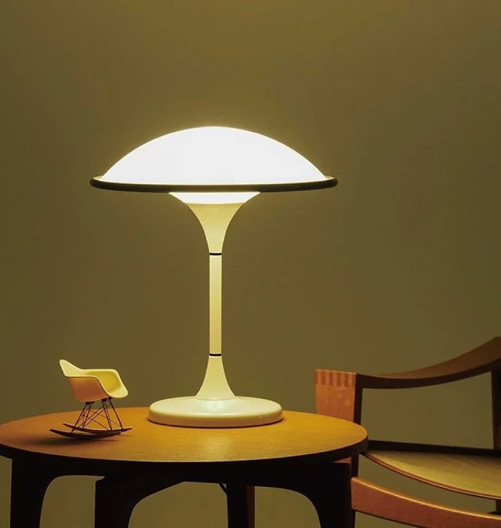 White dome table lamp with a black rim lit up, placed on a round wooden table next to a miniature rocking chair.