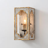A wall-mounted lantern-style sconce with a distressed gold and white finish, featuring an ornate frame around a central glass panel and a single candle-style bulb.
