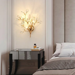 A gold wall sconce with clear crystal accents, emitting light against a cream-colored wall in a bedroom setting.