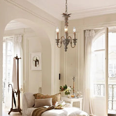Distressed wood candelabra pendant light hanging in a bright, airy living room with large windows, sheer curtains, and a chaise lounge.