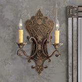 A distressed bronze wall sconce with intricate floral and scroll details, featuring two candle-style lights, mounted on a textured gray wall next to a mirror.