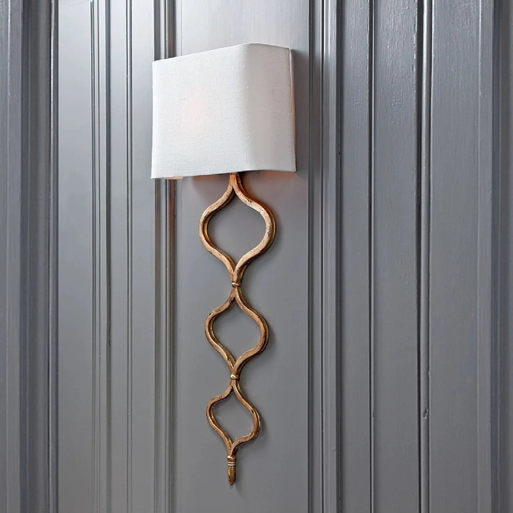 A gold wall sconce with a quatrefoil chain and fabric shade mounted on a dark gray paneled wall.